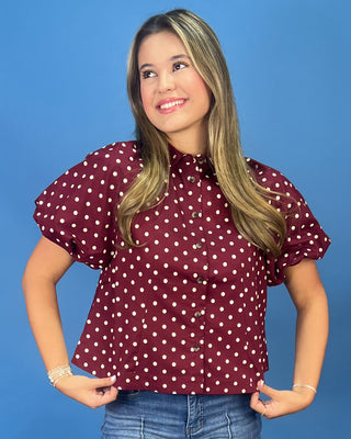A woman modeling a short-sleeve button-up blouse with puff sleeves and a white polka dot pattern on a burgundy base, paired with blue jeans against a solid blue studio background.