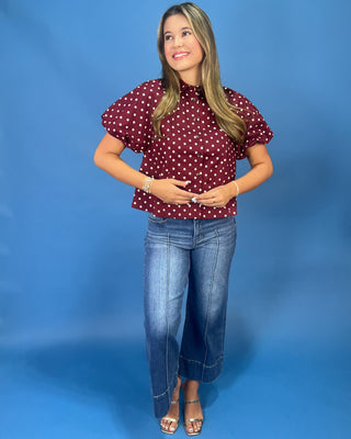 A woman modeling a burgundy short-sleeve blouse with white polka dots, paired with blue jeans and metallic sandals, standing against a solid blue studio backdrop.