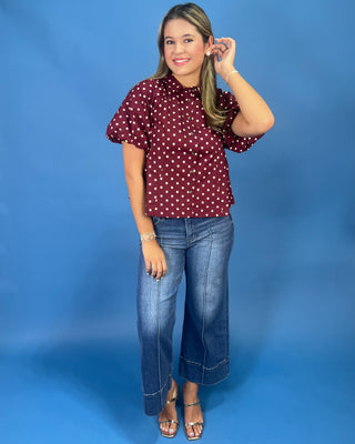 A woman modeling a short-sleeve button-up blouse with a burgundy and white polka dot pattern, paired with blue jeans and metallic sandals, set against a solid blue studio background.