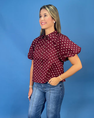 A woman modeling a short-sleeve button-up blouse with puff sleeves, featuring a burgundy red and white polka dot pattern, paired with blue jeans against a solid blue studio background.