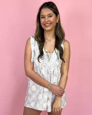 Smiling woman standing against a pink backdrop wearing a sleeveless white floral romper with tie straps, a lace-up V-neck detail, and side pockets, styled with neutral double-strap sandals.