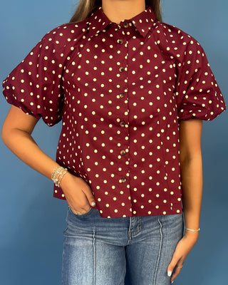 A woman wearing a burgundy short-sleeve button-up blouse with a cream polka dot pattern, paired with blue jeans, set against a solid blue background.