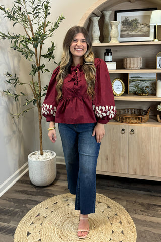 Woman in a burgundy top with white patterns and blue jeans standing in a room with a plant and shelves.