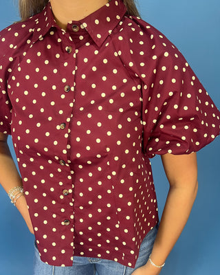 A woman wearing a short-sleeve button-up shirt with a burgundy base and cream polka dots, paired with blue jeans, set against a solid blue background.