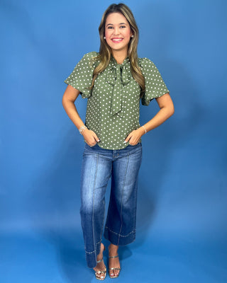 A woman modeling a short-sleeve green blouse with white polka dots and a ruffled tie neckline, paired with blue jeans and metallic sandals, standing against a solid blue studio backdrop.