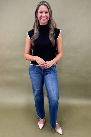 Woman wearing a black top with white trim and blue jeans against a beige background