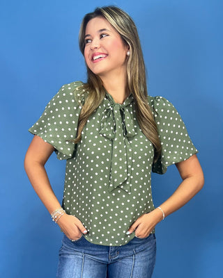 A woman wearing an olive green short-sleeve blouse with white polka dots and a ruffled tie neckline, paired with blue jeans, standing against a solid blue studio background.