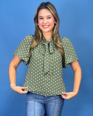 A woman modeling a short-sleeve olive green blouse with white polka dots and a tie-neck detail, paired with blue jeans, against a solid blue studio background.