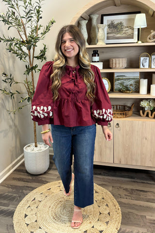 Woman in a red blouse with white patterns standing in a room with wooden flooring and a plant.