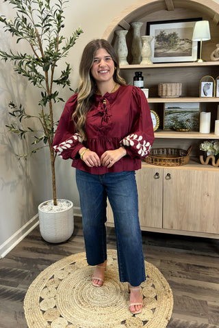 Woman in a burgundy top and blue jeans standing in a room with a plant and shelves.