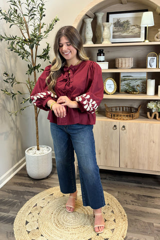 Woman in a burgundy blouse with white patterns and blue jeans standing in a room with wooden shelves and decor.