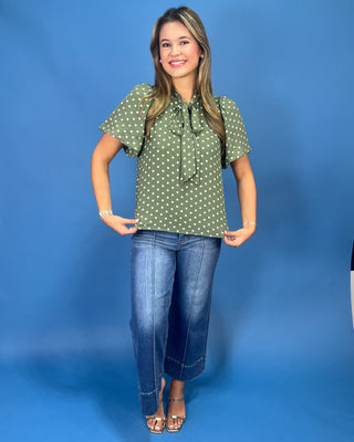 A woman modeling a sage green short-sleeve blouse with white polka dots and a tie-neck detail, paired with blue jeans and metallic sandals, set against a solid blue studio backdrop.