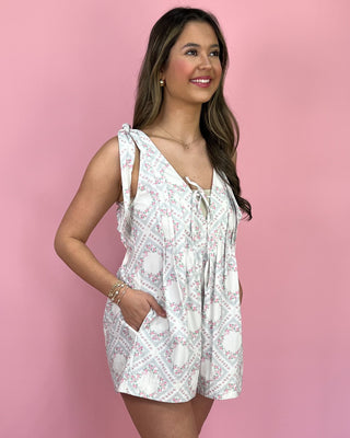 Smiling woman standing against a pink backdrop wearing a sleeveless white floral romper with tie straps, a lace-up V-neck detail, and side pockets, styled with neutral double-strap sandals.