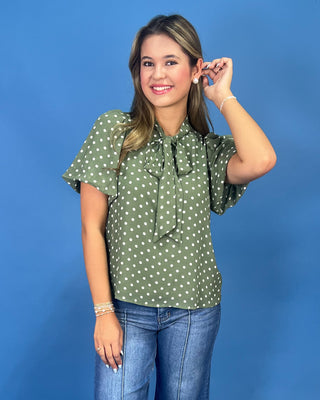 A woman modeling a sage green short-sleeve blouse with white polka dots and a tie-neck bow, paired with blue jeans, against a solid blue studio background.