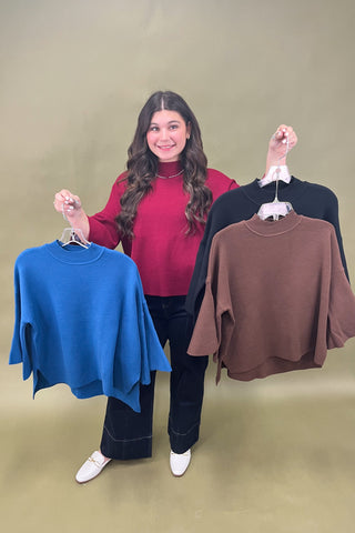 Woman holding two sweaters, one blue and one brown, on hangers against a plain background.