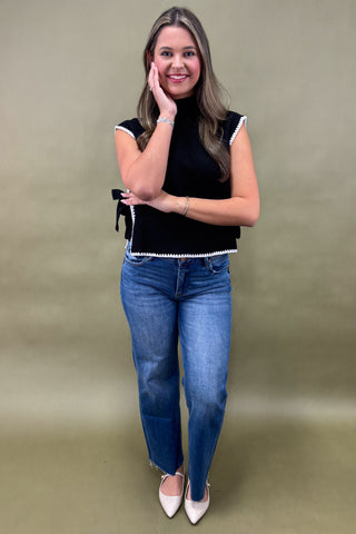 Woman wearing a black top with white trim and blue jeans against a beige background