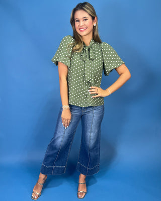 A woman modeling a green short-sleeve blouse with white polka dots, paired with wide-leg blue jeans and metallic heeled sandals, standing against a solid blue studio backdrop.