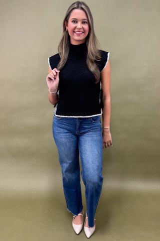 Woman wearing a black sleeveless top with white trim and blue jeans on a beige background