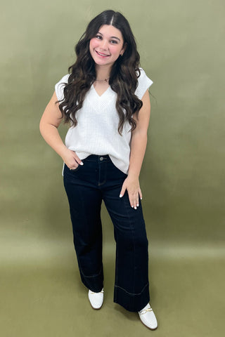 Woman wearing a white sleeveless top and dark blue jeans against a green background