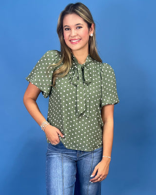 A woman modeling a sage green short-sleeve blouse with white polka dots and a ruffled tie-neck detail, paired with blue jeans, against a solid blue studio background.