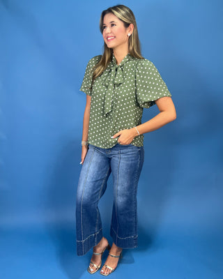 A woman modeling a green short-sleeve blouse with white polka dots and a tie-neck ruffle, paired with blue cropped jeans and metallic sandals, set against a solid blue studio backdrop.