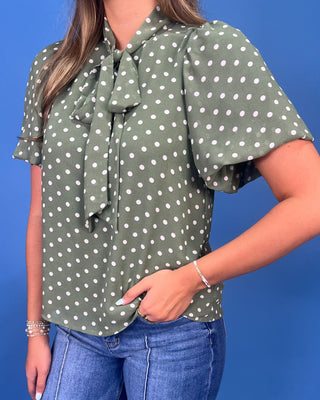 A woman wearing an olive green short-sleeve blouse with white polka dots and a large bow tie at the collar, paired with blue jeans, set against a solid blue background.