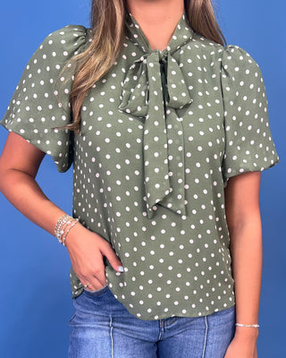 A woman wearing an olive green short-sleeve blouse with white polka dots and a large front bow tie, paired with blue jeans against a solid blue background.