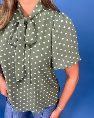 A woman wearing a sage green short-sleeve blouse with white polka dots and a large bow tie neckline, paired with blue jeans against a solid blue background.