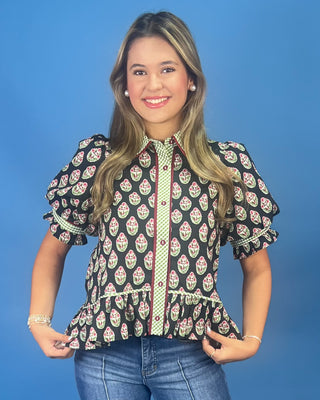 A woman modeling a short-sleeve button-up blouse with a black background and a colorful paisley-like print in pink, green, and white, paired with blue jeans against a solid blue studio background.