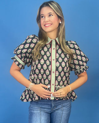 A woman modeling a black short-sleeve blouse with a colorful paisley block print and a green-and-white patterned placket, paired with blue jeans against a solid blue background.