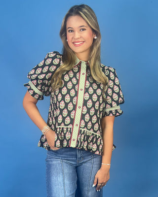 A woman modeling a black short-sleeve blouse with a green and pink paisley pattern, ruffled sleeves and hem, and a green polka-dot collar, paired with blue jeans against a solid blue background.