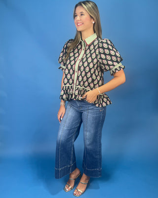 A woman modeling a short-sleeve button-up blouse with a black base and a colorful paisley pattern, paired with blue jeans and metallic sandals, set against a solid blue studio background.