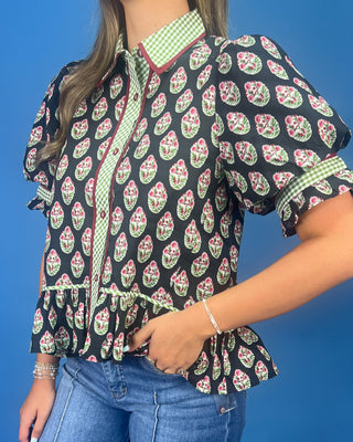 A woman wearing a black short-sleeve blouse with a pink and green paisley print, a green and white gingham collar, and a ruffled hem, paired with blue jeans against a solid blue background.