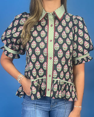 A woman wearing a short-sleeve button-up blouse with a black background, green and white gingham trim, and a pink and green floral block print, paired with blue jeans against a solid blue background.
