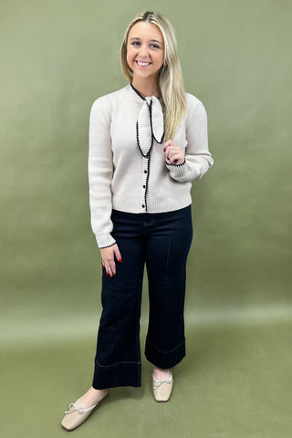 Woman wearing a light-colored cardigan with black buttons and dark pants on a green background