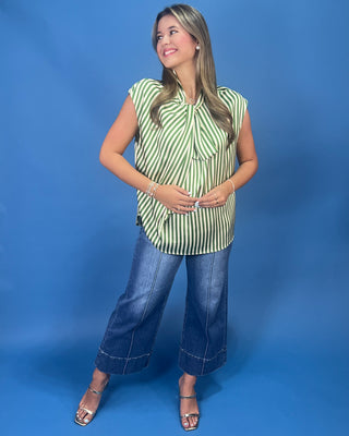 A woman modeling a sleeveless green and white striped blouse with a tie-neck detail, paired with wide-leg blue jeans and metallic sandals, set against a solid blue studio backdrop.