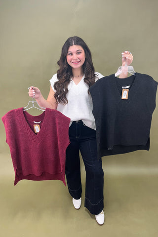 Woman holding a maroon and a black sleeveless sweater against a green background