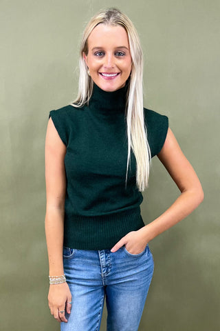 Woman wearing a dark green sleeveless sweater and blue jeans against a plain background