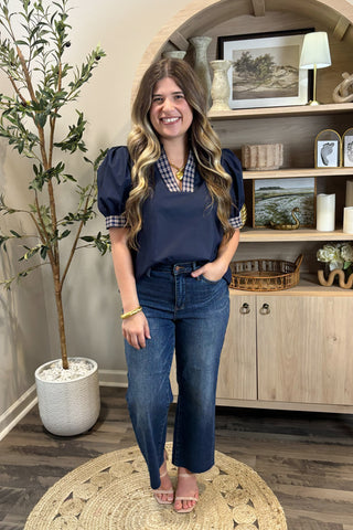 Woman in navy top and jeans standing in a room with a plant and wooden cabinet.