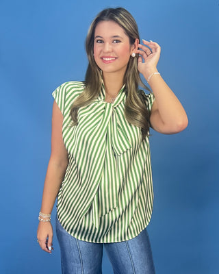 A woman modeling a sleeveless top with green and white vertical stripes and a tie-neck detail, paired with blue jeans, against a solid blue studio background.