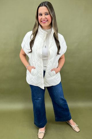 Woman wearing a white puffer vest over a white shirt and blue jeans on a green background