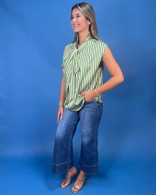 A woman modeling a sleeveless green and white striped blouse with a tie-neck detail, paired with blue cropped jeans and metallic sandals, set against a solid blue studio backdrop.
