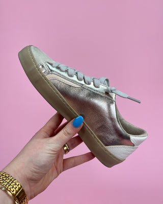 Silver sneaker held by a hand with blue nail polish against a pink background