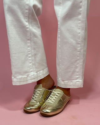 Gold sneakers worn with white pants on a pink background