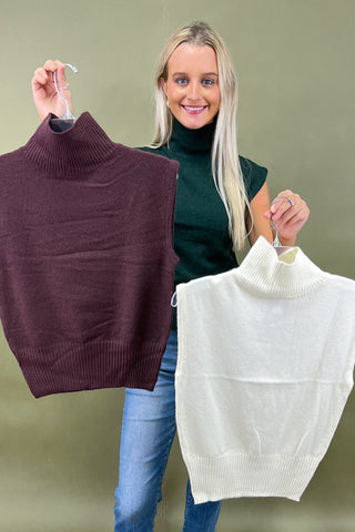 Woman holding two sweater vests, one brown and one white, against a plain background.