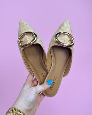 Pair of beige pointed-toe flats with gold circular details held against a pink background