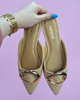 Gold high-heeled shoes with a decorative ring held by a hand against a purple background