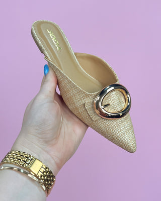 Beige pointed-toe shoe with gold buckle held by a hand against a pink background