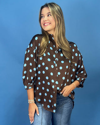 A woman wearing a brown button-up blouse with light blue polka dots, paired with blue jeans, standing against a solid blue background.