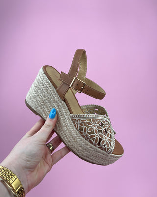 Brown wedge sandal with woven sole held by a hand against a pink background
